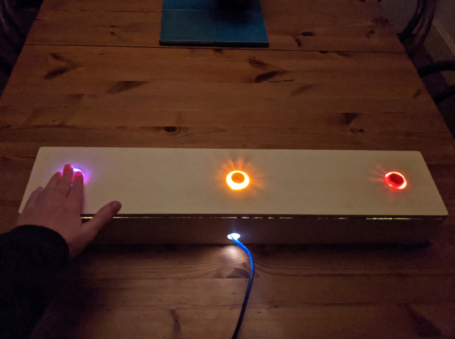 A photo of a very wide wooden box with 3 arcade buttons spaced along it and a hand pressing the left button.