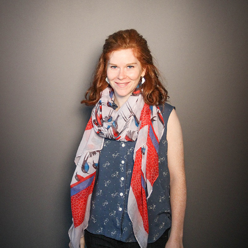 A photo of me; I have longish ginger hair and am wearing a blue shirt covered in bicycles, and a big summer scarf with birds on it.